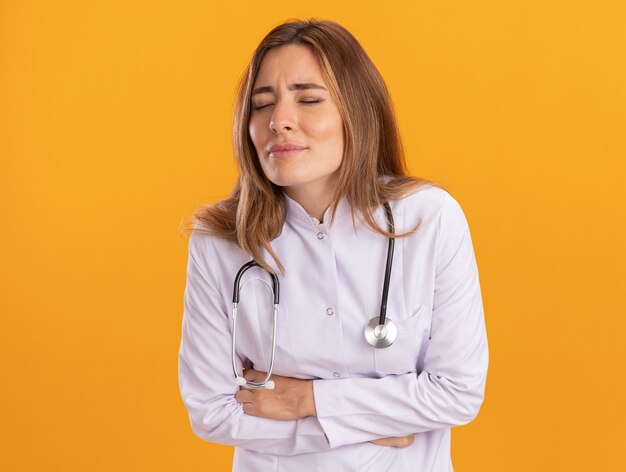 Mal aux yeux fermés jeune femme médecin portant une robe médicale avec stéthoscope attrapé estomac douloureux isolé sur mur jaune