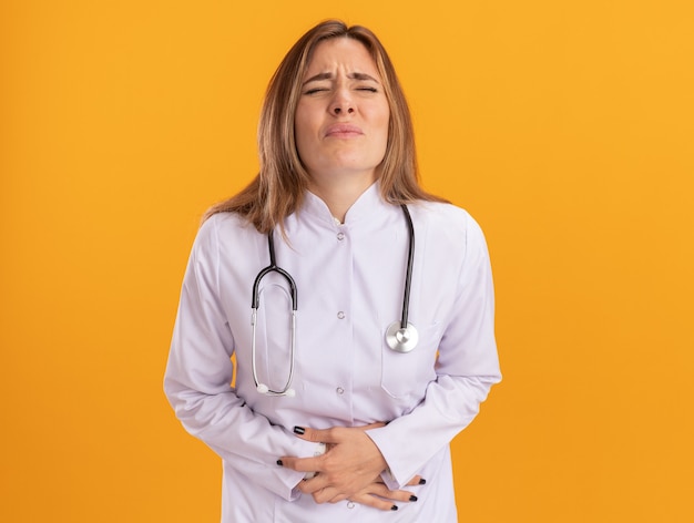 Mal aux yeux fermés jeune femme médecin portant une robe médicale avec stéthoscope saisi estomac isolé sur mur jaune