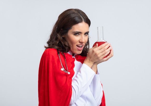 Photo gratuite mal superwoman joyeuse en uniforme de médecin avec cape rouge et stéthoscope détient un liquide chimique rouge dans un flacon de verre isolé sur un mur blanc