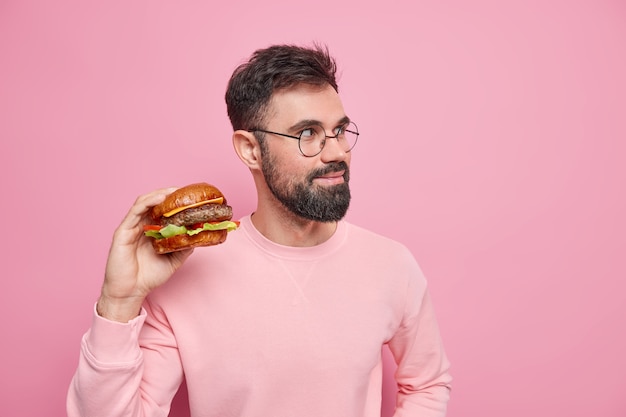 Malbouffe et concept de nutrition malsaine. L'homme adulte barbu tient un délicieux hamburger a une collation rapide regarde pensivement