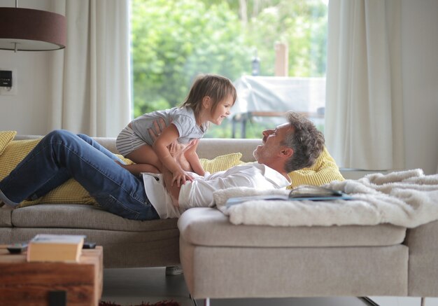 Mâle adulte allongé sur le canapé et jouant avec son enfant sous la lumière du soleil à travers les fenêtres