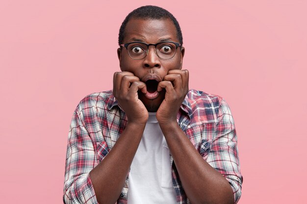 Mâle aux yeux écorchés à la peau foncée émotionnelle dans les lunettes, garde la bouche largement ouverte, se sent nerveusement devant quelque chose, vêtu d'une chemise à carreaux, isolé sur rose