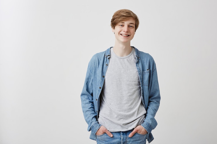 Photo gratuit le mâle blond clair aux yeux bleus et aux croisillons sourit doucement comme étant heureux après la date avec un ami. un adolescent qui réussit est heureux d'entendre de bonnes nouvelles et tient les mains dans des poches de jeans