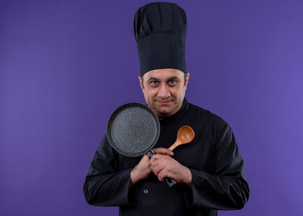 Photo gratuite mâle chef cuisinier en uniforme noir et chapeau de cuisinier tenant une poêle et une cuillère en bois croisant les mains regardant la caméra en souriant debout sur fond violet