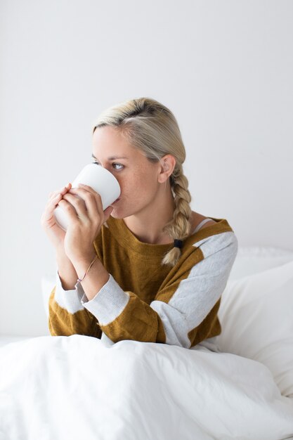 Malheureuse femme assise sur le lit et buvant une boisson chaude