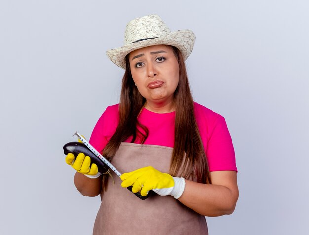 Malheureuse femme jardinier d'âge moyen en tablier et chapeau portant des gants en caoutchouc tenant des aubergines fraîches en le mesurant avec un ruban à mesurer regardant la caméra avec une expression triste debout sur fond blanc