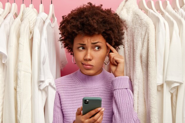 Malheureuse femme à la peau sombre regarde à travers une armoire, se tient entre des tenues blanches sur des cintres, garde l'index sur la tempe, tient un téléphone mobile moderne