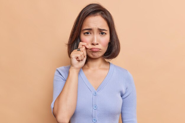 Malheureuse frustrée jeune femme asiatique sanglote et frotte les larmes se sent désespérée porte un pull bleu a une expression insatisfaite de la chaleur brisée isolée sur un mur beige