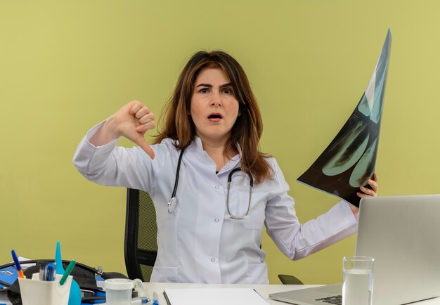 Malheureux femme médecin d'âge moyen portant une robe médicale et un stéthoscope assis au bureau avec des outils médicaux et un ordinateur portable tenant un tir aux rayons x montrant le pouce vers le bas isolé