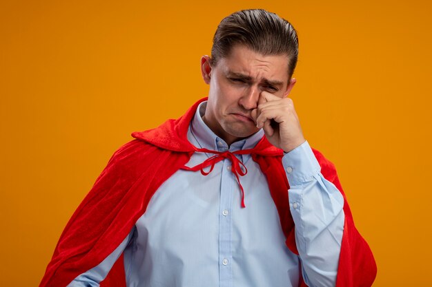 Malheureux homme d'affaires de super héros en cape rouge pleurer frottant les yeux debout sur le mur orange