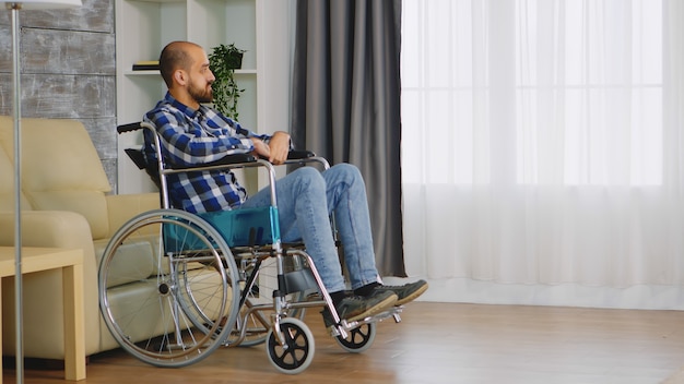 Malheureux homme en fauteuil roulant dans le salon en regardant la fenêtre.