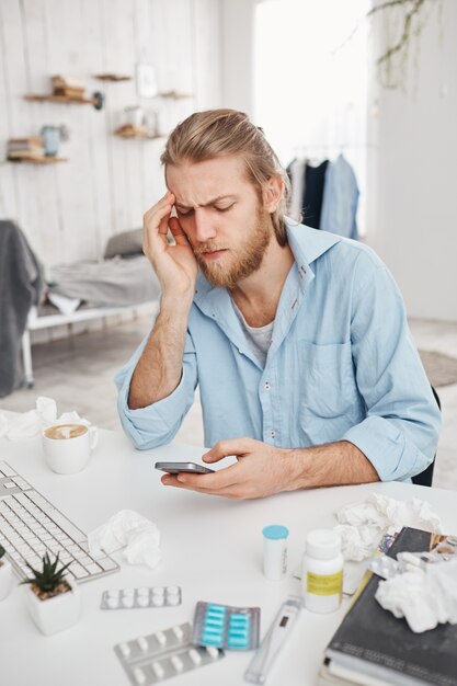 Malheureux jeune homme barbu gestionnaire assis à table entouré de pilules, comprimés, médicaments. Un employé de bureau blond a un mauvais rhume, surfe sur Internet, souffre de températures élevées. Problèmes de santé.