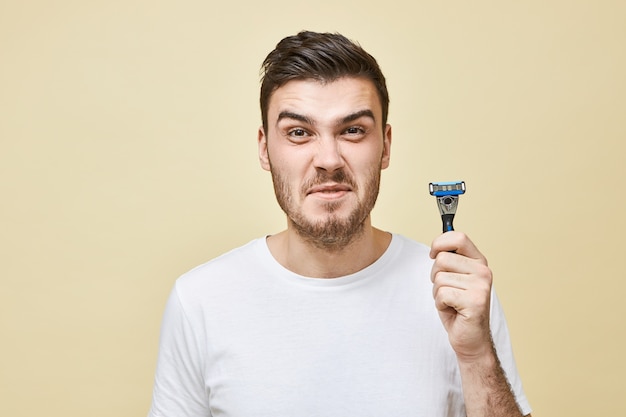 malheureux mécontent jeune homme brune avec des soies grimaçant ne veut pas se raser la barbe, détestant le processus de rasage, ayant la peau sensible, posant isolé avec un rasoir dans les mains