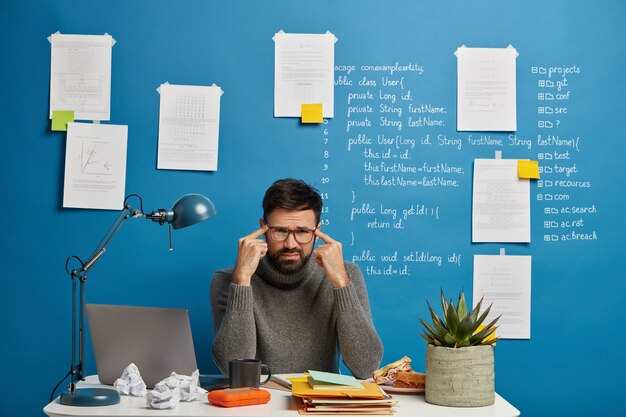 Malheureux travailleur masculin dans les lunettes est assis au bureau pendant la dure journée de travail, garde les doigts sur les tempes, souffre de maux de tête, essaie de se concentrer sur l'objet, fatigué de la surcharge de travail à l'ordinateur portable
