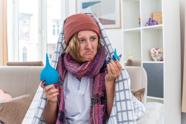 Malsaine jeune femme au chapeau enveloppé dans une couverture se sentir mal et malade tenant des lavements à la confusion d'avoir des doutes assis sur un canapé dans un salon lumineux