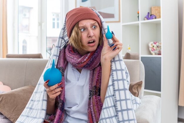 Malsaine jeune femme au chapeau enveloppé dans une couverture se sentir mal et malade tenant des lavements à la confusion d'avoir des doutes assis sur un canapé dans un salon lumineux