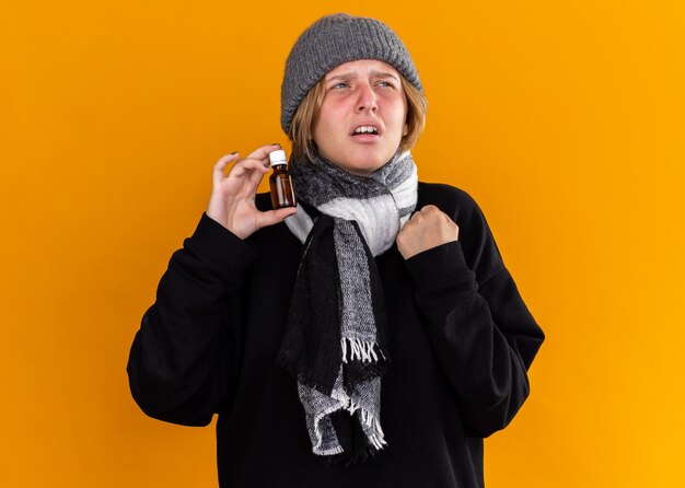 Malsaine jeune femme portant un chapeau chaud et avec un foulard autour de son cou se sentir malade souffrant du rhume et de la grippe tenant la bouteille de médicaments à côté avec une expression agacée sur le mur orange