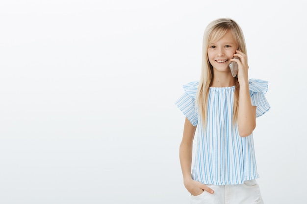 Maman a donné un téléphone portable à sa fille pour parler avec sa grand-mère. Heureux enfant européen aux cheveux blonds en blouse bleue, debout avec désinvolture sur un mur gris et communiquant via smartphone