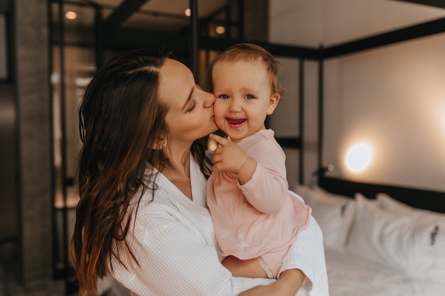 Maman embrasse l'enfant blond regardant la caméra avec le sourire. Femme tient sa fille dans ses bras sur fond de lit blanc.