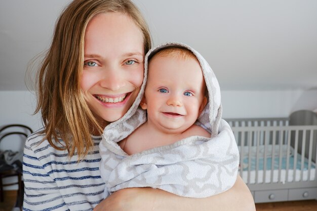 Maman joyeuse tenant bébé heureux portant une serviette à capuchon après le bain ou la douche. Vue de face. Concept de garde d'enfants ou de bain