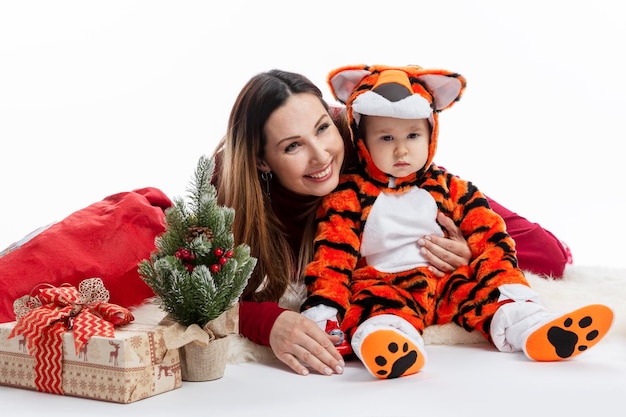 Maman avec un petit fils avec un arbre de noël et des cadeaux. une jeune femme souriante avec un bébé dans un costume de fourrure de tigre - le symbole de 2022. fond blanc. vacances d'hiver traditionnelles.