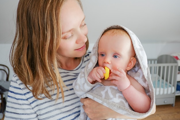 Maman positive tenant doux bébé sec portant une serviette à capuche après la douche et mordant le jouet de bain en caoutchouc Vue de face. Concept de garde d'enfants ou de bain