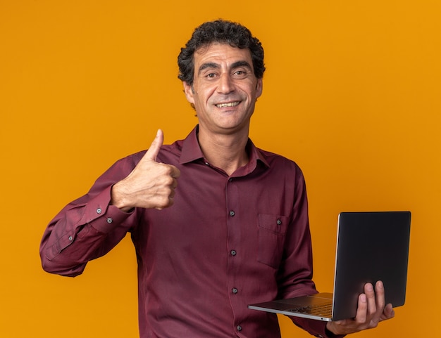 Man in purple shirt holding laptop looking at camera smiling confiant showing Thumbs up debout sur fond orange