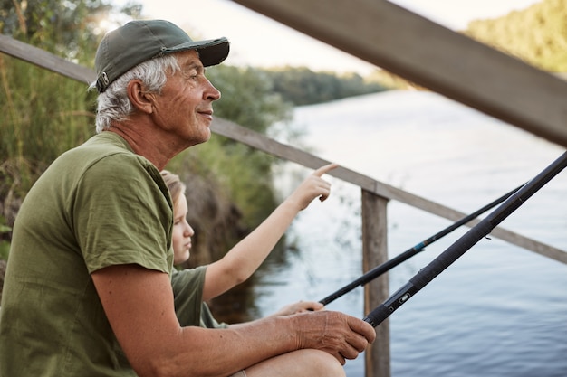 Man avec son petit-fils assis sur un ponton en bois avec des cannes à pêche dans les mains, profitant de la belle nature, petit garçon pointant quelque chose avec son doigt.