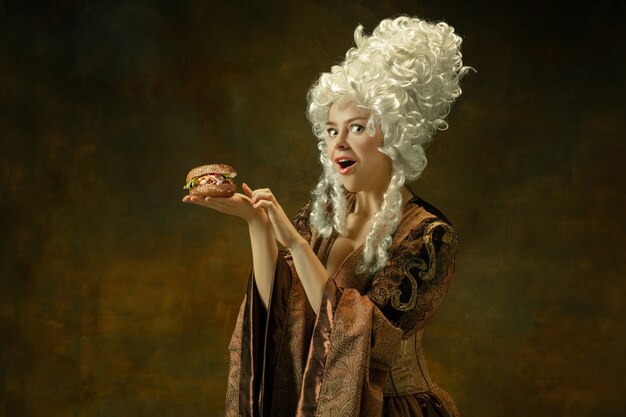 Manger un hamburger se demanda. Portrait de jeune femme médiévale en vêtements vintage marron sur fond sombre. Modèle féminin en tant que duchesse, personne royale. Concept de comparaison des époques, moderne, mode, beauté.