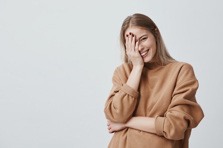 Photo gratuit la mannequin femme adorable positive sourit joyeusement avec de longs cheveux blonds, heureuse de recevoir des nouvelles agréables, garde la main sur la tête, porte des vêtements amples beiges. concept de personnes, de style et d'émotions.