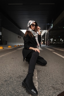 Mannequin jeune femme en lunettes de soleil élégantes en foulard élégant en soie sur la tête en manteau noir tendance en jeans pour jeunes en bottes posant assis sur l'asphalte dans la ville. fille d'affaires se détend sur l'asphalte.