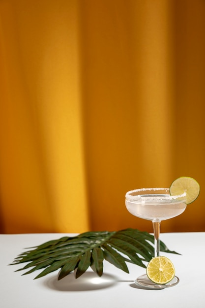 Margarita cocktail avec des limes en tranches et des feuilles de palmier sur une table blanche près de rideau jaune