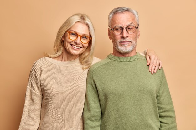 Mari et femme âgés posent pour le portrait de famille embrasser sourire positivement vêtus de lunettes cavaliers debout contre le mur de studio brun