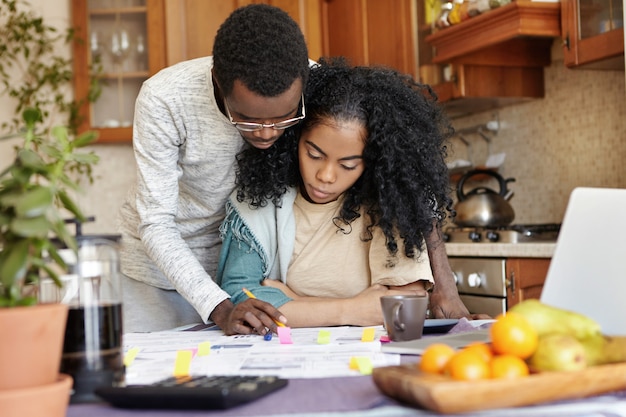 Mari à lunettes aidant sa belle femme avec la paperasse, debout à côté d'elle et expliquant quelque chose sur des papiers. Jeune famille africaine gérant les finances ensemble, assis à la table de la cuisine