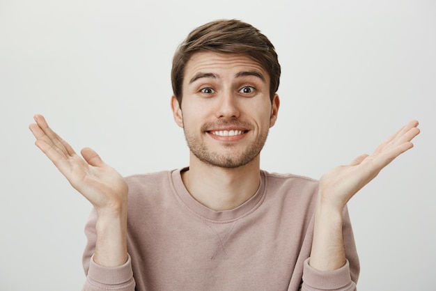 Le mari n'a aucune idée Portrait intérieur d'un beau jeune homme avec des poils haussant les épaules et levant les paumes souriant maladroitement comme s'il s'excusait debout sur fond gris confus