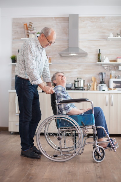 Mari regardant une femme âgée handicapée dans la cuisine. Femme âgée handicapée assise en fauteuil roulant dans la cuisine en regardant par la fenêtre. Vivre avec une personne handicapée. Mari aidant sa femme avec un handicap