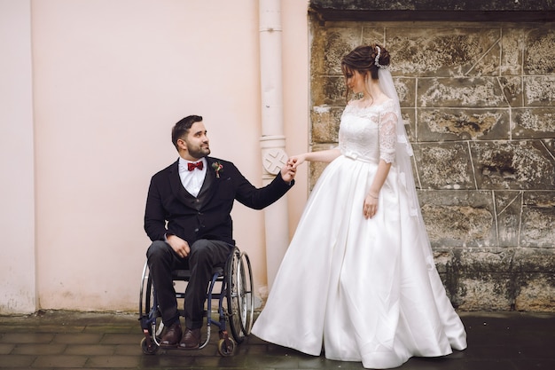 Marié sur le fauteuil roulant tient la main de la mariée debout devant la vieille maison dans la rue