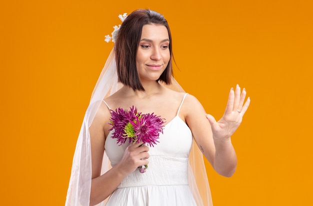 Mariée en belle robe de mariée avec bouquet de fleurs en regardant sa bague à son doigt debout sur un mur orange