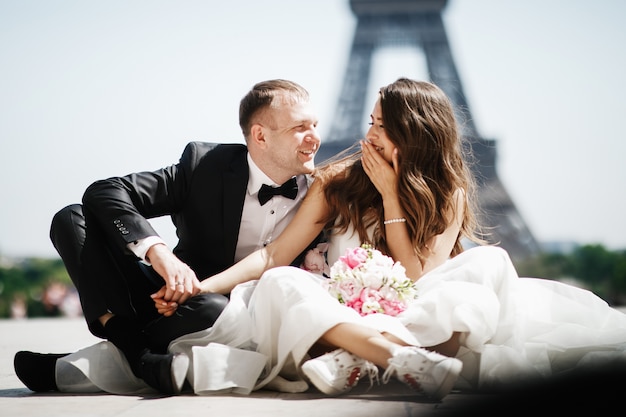 Photo gratuite la mariée est assise dans des ricanements devant la tour eiffel à paris tandis que le marié tient sa main