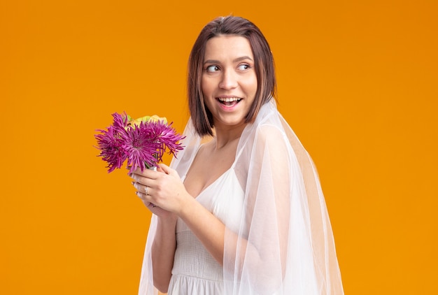 Mariée heureuse et joyeuse dans une belle robe de mariée va jeter un bouquet de fleurs de mariage souriant confiant debout sur un mur orange