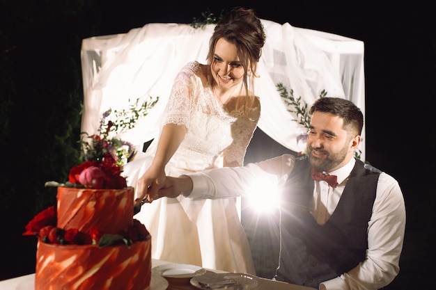 Mariée et le marié dégustent gâteau de mariage rouge debout devant l&#39;autel dans la nuit