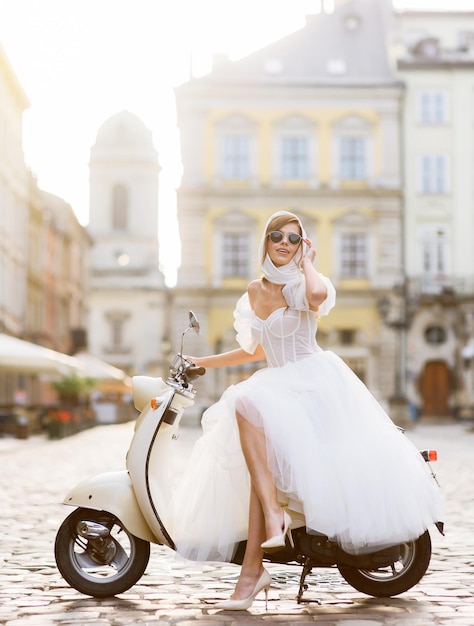 Mariée en robe de mariée et lunettes de soleil posant sur un scooter