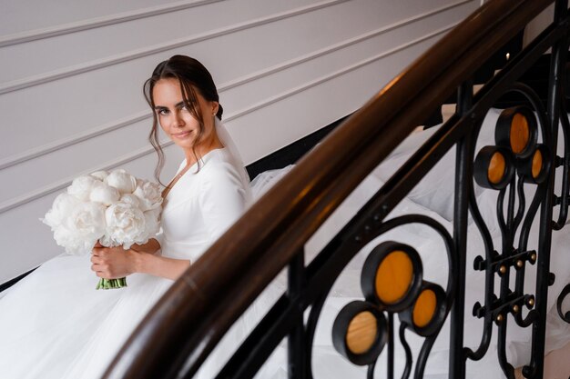Mariée romantique en robe de satin classique tenant un bouquet pf pionies en remuant dans les escaliers à l'intérieur La femme a une belle coiffure de mariage avec des accessoires de voile et un maquillage naturel nude