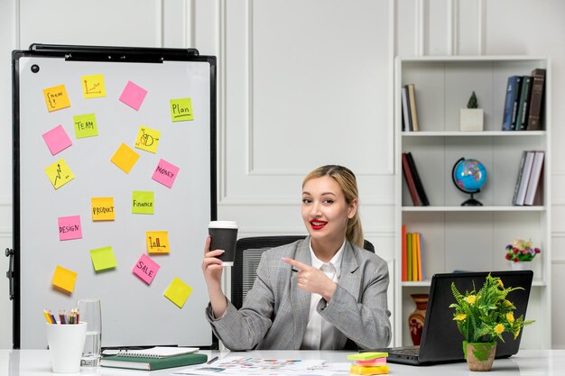 Marketing jeune jolie fille blonde en costume gris au bureau pointant vers une tasse en papier