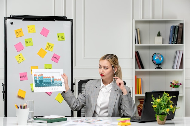 Photo gratuite marketing jeune jolie fille blonde en costume gris au bureau tenant un stylo et des statistiques