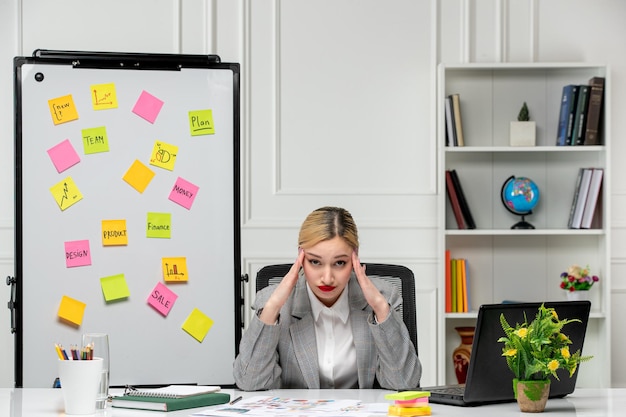 Marketing jolie jeune fille blonde en costume gris au bureau fatiguée et confuse