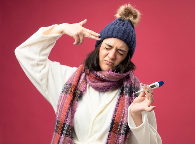 Photo gratuite marre de jeune fille malade de race blanche portant chapeau d'hiver robe et écharpe tenant un thermomètre faisant le geste de suicide avec les yeux fermés isolé sur le mur cramoisi