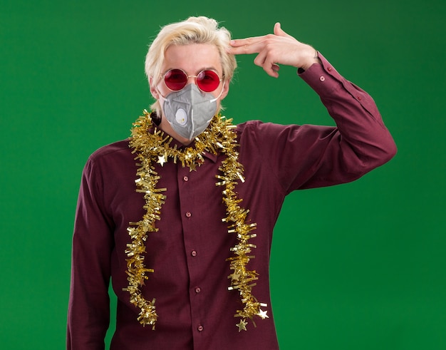 Photo gratuite marre de jeune homme blond portant des lunettes et un masque de protection avec guirlande de guirlandes autour du cou regardant la caméra faisant un geste de suicide isolé sur fond vert