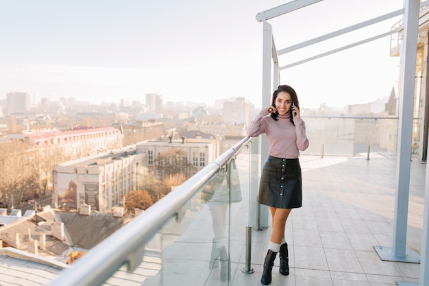 Matin ensoleillé dans la grande ville sur la terrasse de la jeune femme à la mode brune parlant au téléphone et souriant. Jolie femme modèle, refroidissement, temps de détente, bonheur.