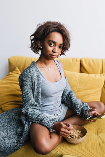 Matin de jeune fille ayant un petit-déjeuner sain, gruau, granola, textos au téléphone, assis sur un canapé jaune à la maison. Elle a les cheveux courts et bouclés, porte un cardigan décontracté gris, un short.